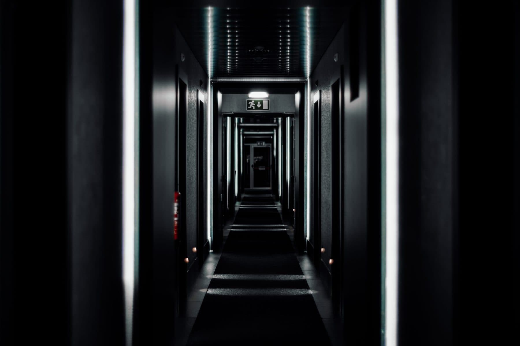 A long, dark, symmetrical corridor with an exit sign at the far end