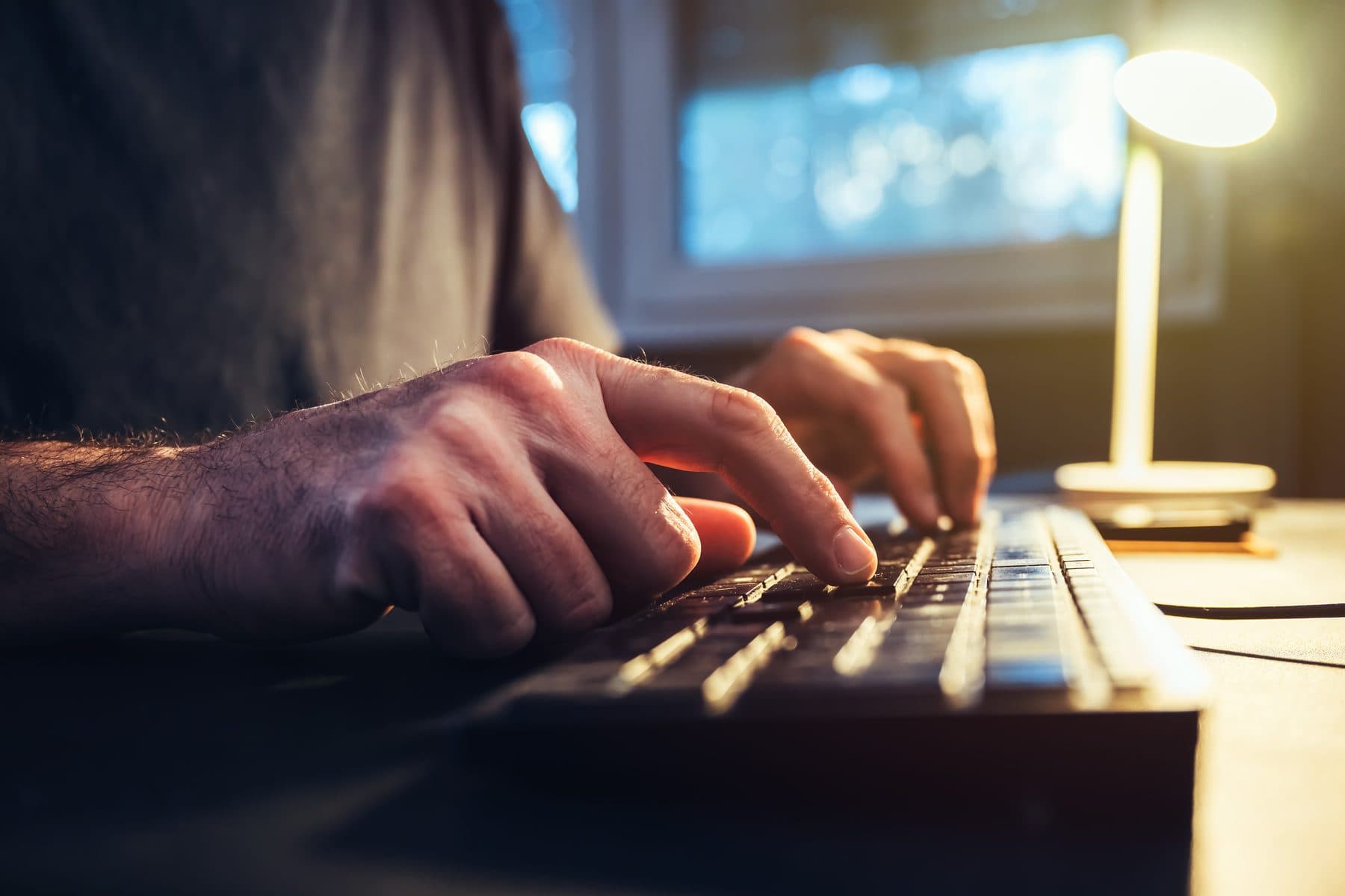 Hands typing on a backlit keyboard under a desk lamp